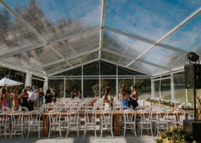 Clear marquee hire with transparent roof and sides set with white chairs, wooden tables and linenwith sun umbrellas and outdoor furniture hire. With people