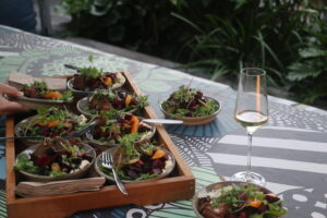 Outdoor table with multiple bowls of colorful salad on a wooden tray and a glass of white wine nearby
